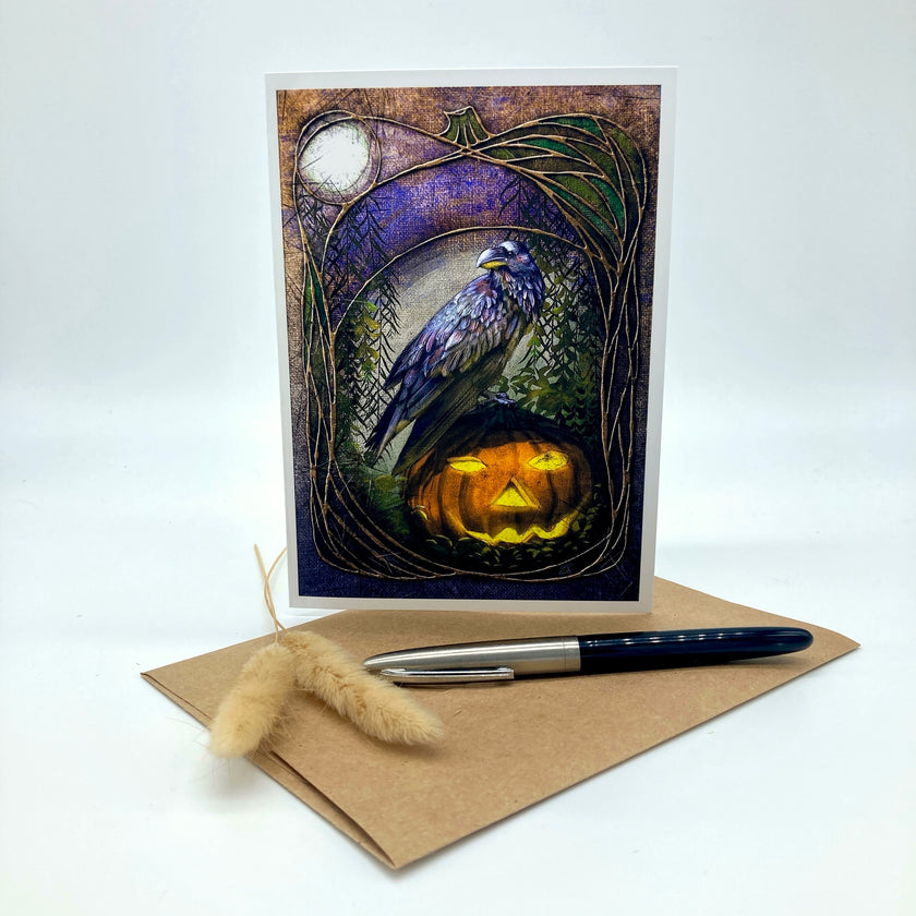 An art print or greeting card featuring a raven perched above a glowing jack-o'-lantern, displayed upright on a kraft envelope with a pen and dried grass stem on a white studio background.