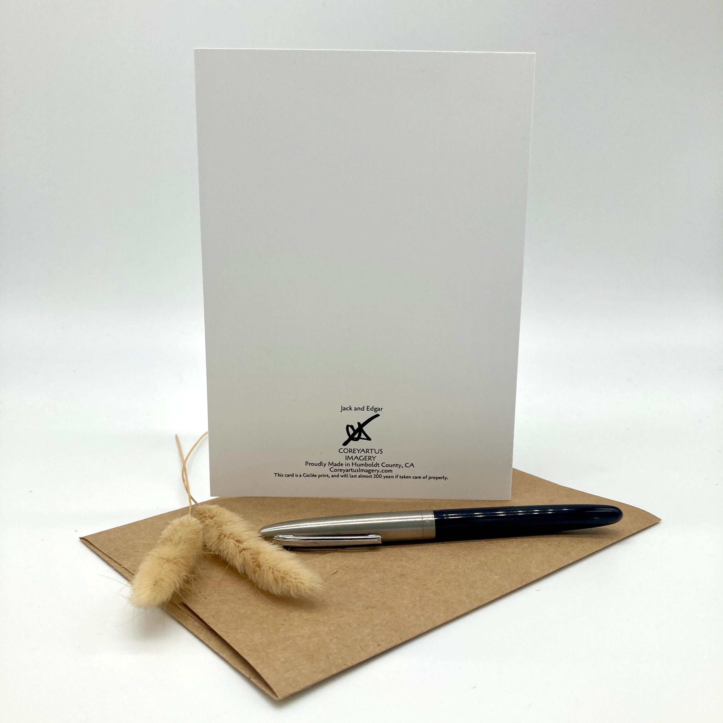 A white stationery card with black printed branding and address stands upright on a brown kraft envelope, with a black and silver pen and a small dried beige grass sprig in front, photographed on a clean white background.
