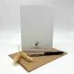 A white stationery card with black printed branding and address stands upright on a brown kraft envelope, with a black and silver pen and a small dried beige grass sprig in front, photographed on a clean white background.