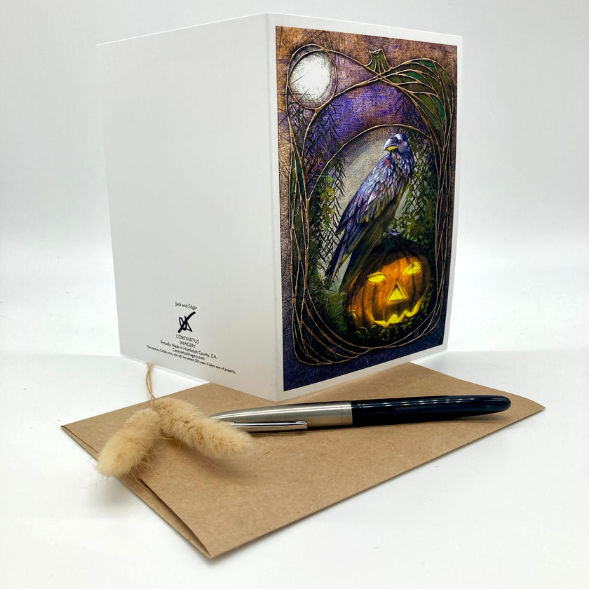 A white greeting card with a Halloween-themed illustration of a raven perched on a glowing jack-o'-lantern, displayed with a kraft envelope and a black-and-silver pen on a white background.