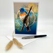 A greeting card featuring a detailed illustration of a bluebird perched on a branch against an abstract blue and gold background. The card is displayed upright on a white surface with an envelope, a black pen, and two dried bunny tails.