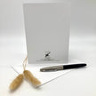 A white greeting card with a black printed logo and text stands upright on a white surface, with an envelope and a black-and-silver pen placed in front. Two small dried grass stems decorate the left side.