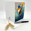 A white greeting card standing open on a white surface, featuring a colorful bird illustration on the front. A black pen rests across a white envelope below the card, with two small dried beige grass stems in the foreground.