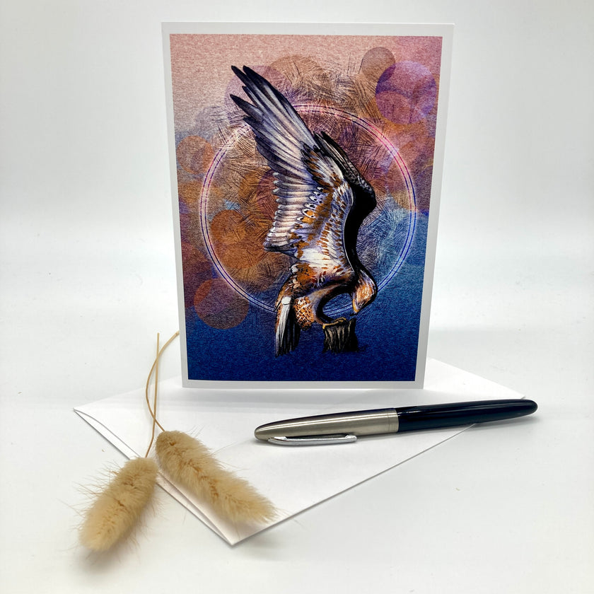 A greeting card featuring a stylized bird with outstretched wings in blue, purple, and orange tones, displayed upright on a white surface with an envelope, a black pen, and two dried beige grass stems.