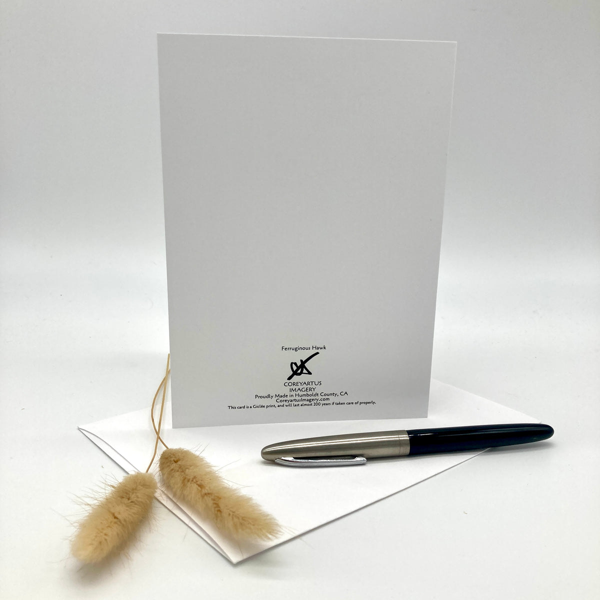 A white stationery card with black printed text and logo at the bottom, displayed upright on a white surface with a white envelope underneath and a black-and-silver pen resting on top. Two small beige dried grass stems are placed in front.
