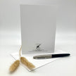 A white stationery card with black printed text and logo at the bottom, displayed upright on a white surface with a white envelope underneath and a black-and-silver pen resting on top. Two small beige dried grass stems are placed in front.