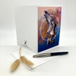 A white greeting card with a colorful illustration of a bird of prey in flight, displayed upright with an envelope and a black pen on a white surface. The card features a white left panel with a small black logo and text, and a vibrant right panel with a blue, purple, and orange background and a detailed bird design.