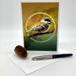 A vertical greeting card featuring a detailed illustration of a chickadee bird perched on a branch against a warm brown and green background, displayed on a white surface with a pinecone and a black-and-silver pen.