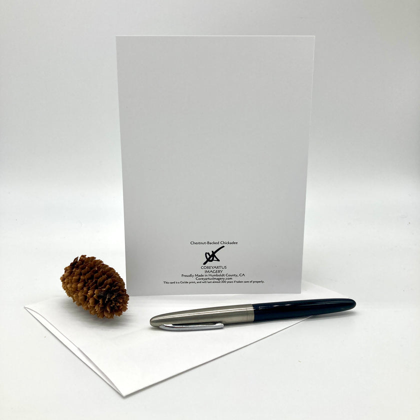 A white stationery set on a light gray background, featuring a vertical white card with small black text and a pinecone, alongside a black and silver pen resting on a folded white sheet.