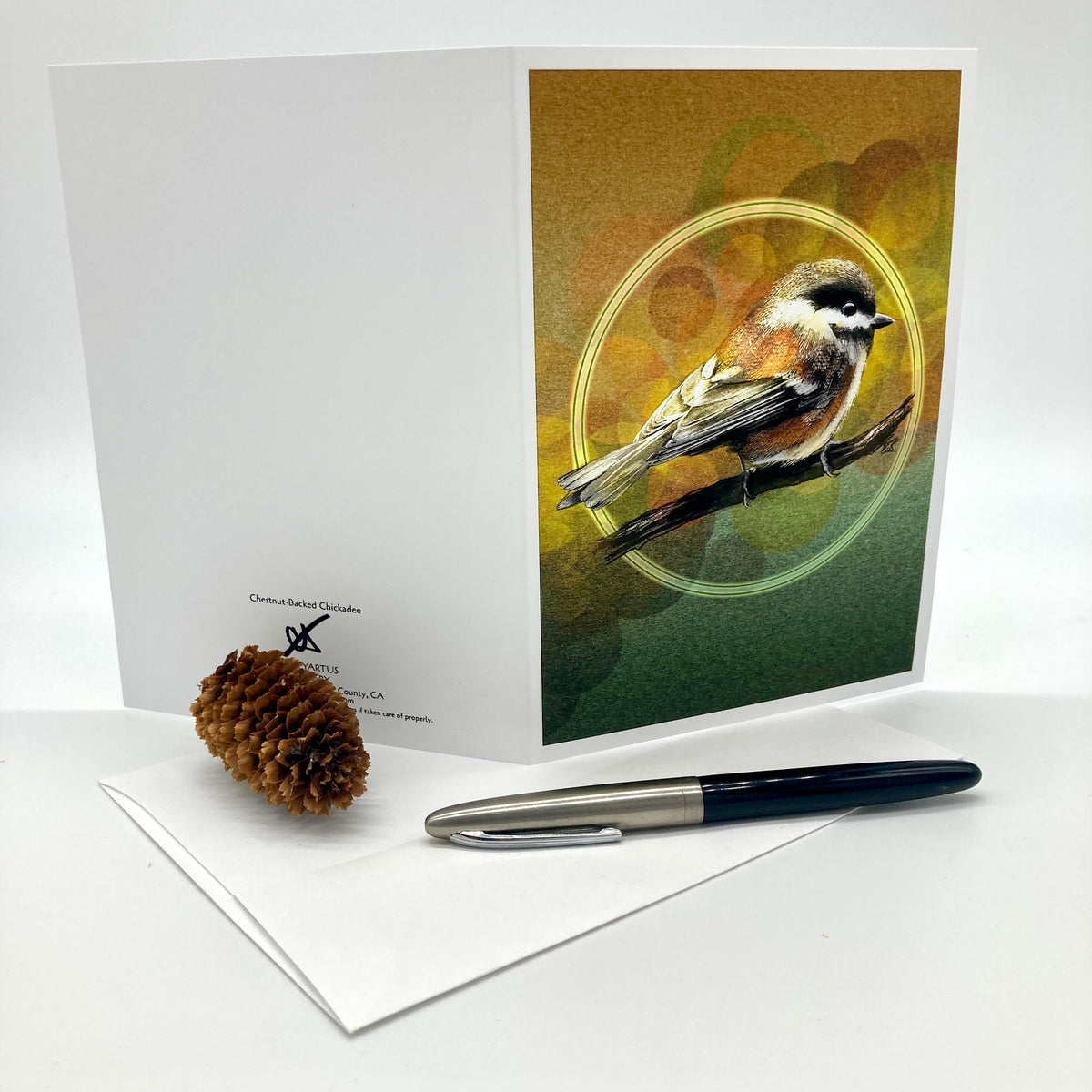 A greeting card with a printed illustration of a small bird perched on a branch against an abstract gold and green background, displayed upright with a white envelope, a pinecone, and a pen on a white surface.