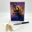A vertical greeting card featuring a colorful bird wearing a top hat and a monocle perched on a branch, displayed upright on a white surface with an envelope and a black-and-silver pen nearby.