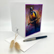 A white greeting card standing upright on a white surface, featuring a colorful illustration of a bird wearing a top hat on the front. A white envelope and a black-and-silver pen are placed in front, with dried grass stems to the left.