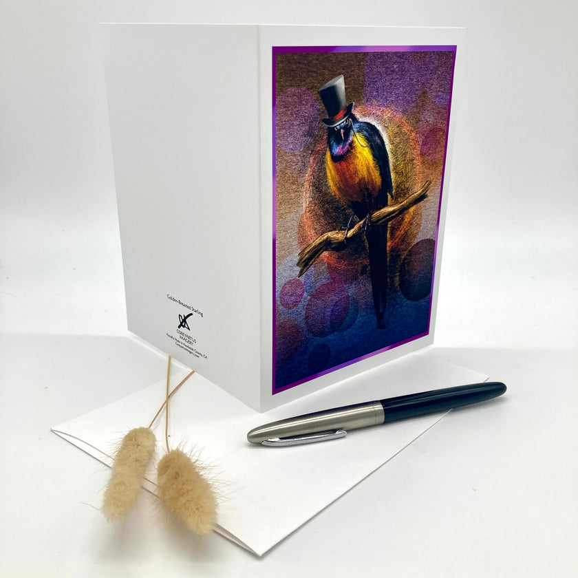 A white greeting card standing upright on a white surface, featuring a colorful illustration of a bird wearing a top hat on the front. A white envelope and a black-and-silver pen are placed in front, with dried grass stems to the left.