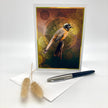 A greeting card featuring a bird illustration, displayed upright on a white surface with a pen and dried bunny tail stems as props.