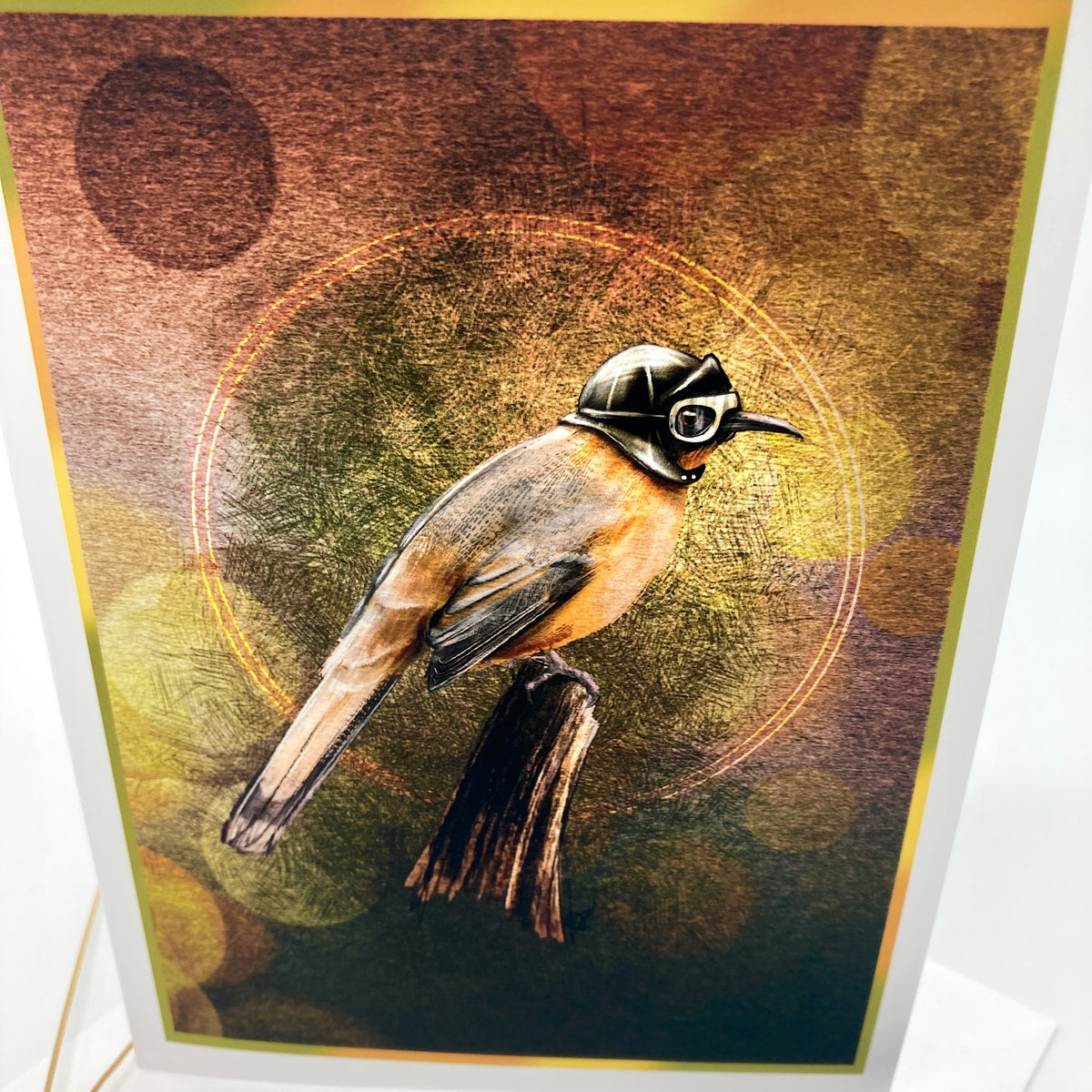 An art print or card featuring a bird wearing a small black helmet and goggles, perched on a branch against a textured abstract background with warm brown, gold, and olive tones. The print is photographed at a slight angle with a white border visible around the artwork.