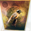 An art print or card featuring a bird wearing a small black helmet and goggles, perched on a branch against a textured abstract background with warm brown, gold, and olive tones. The print is photographed at a slight angle with a white border visible around the artwork.