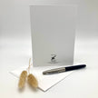 A white greeting card with a black printed logo for 'Ruben Silva Card Company' stands upright on a white surface, with a black and silver pen resting on a white envelope in front and dried beige grass stems to the left.