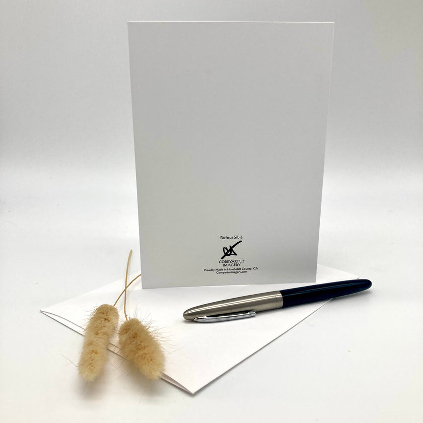 A white greeting card with a black printed logo for 'Ruben Silva Card Company' stands upright on a white surface, with a black and silver pen resting on a white envelope in front and dried beige grass stems to the left.