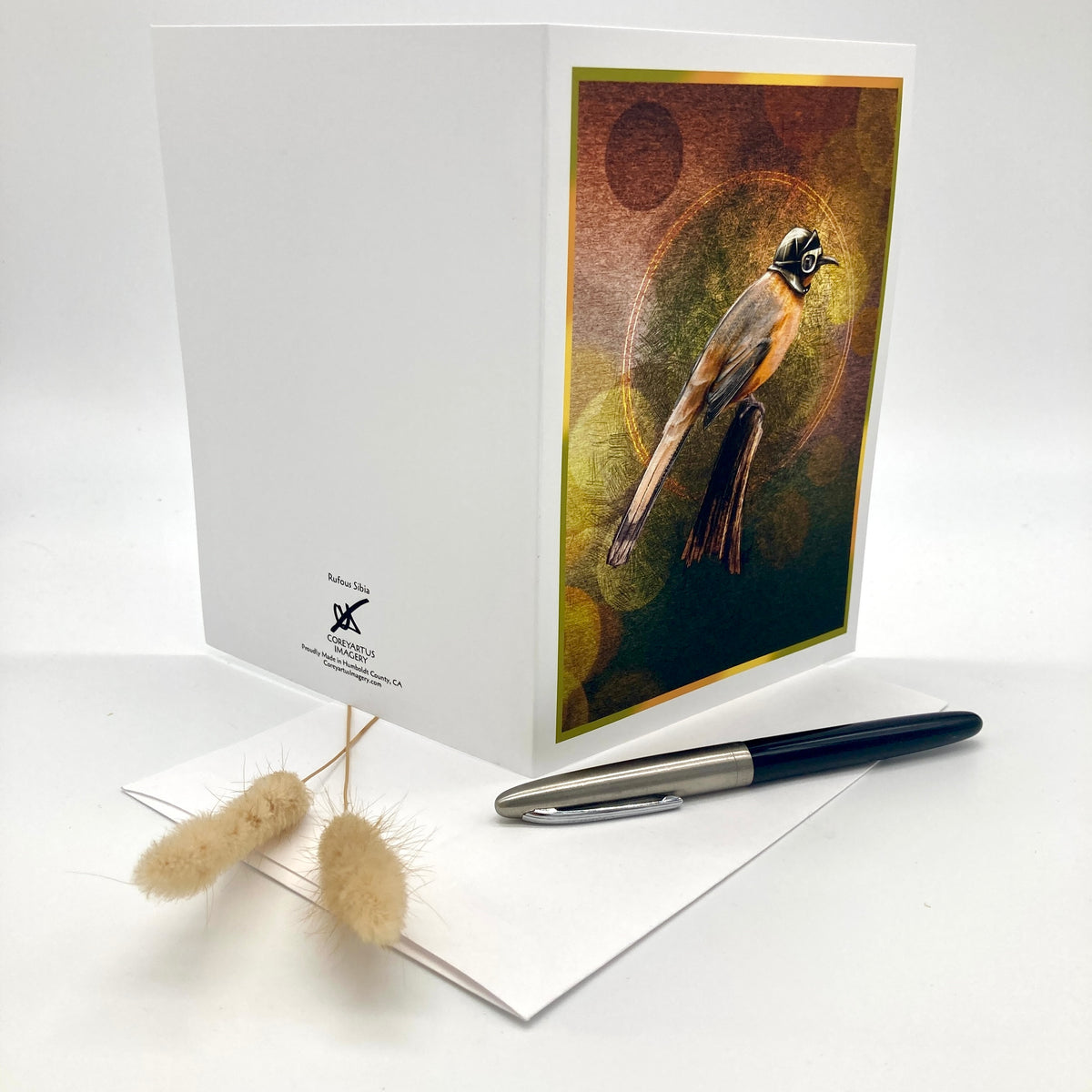 A white greeting card with a printed illustration of a bird on the front cover, displayed upright on a white surface with a pen and dried grass stems beside it.