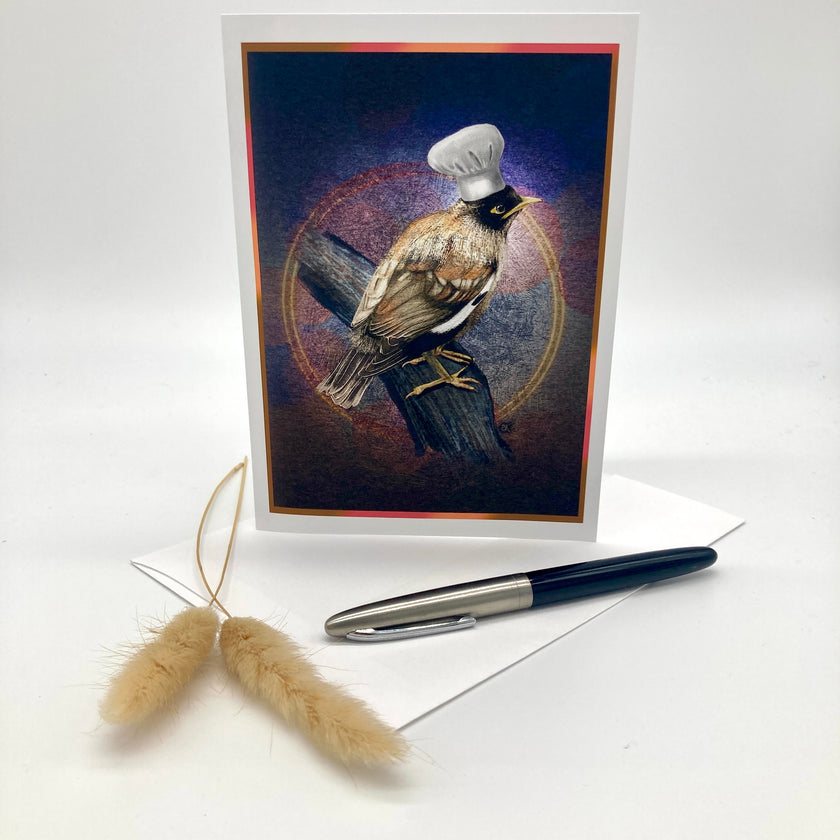 A greeting card featuring an illustration of a bird wearing a chef's hat, displayed upright on a white surface with an envelope, a pen, and two dried bunny tail stems.