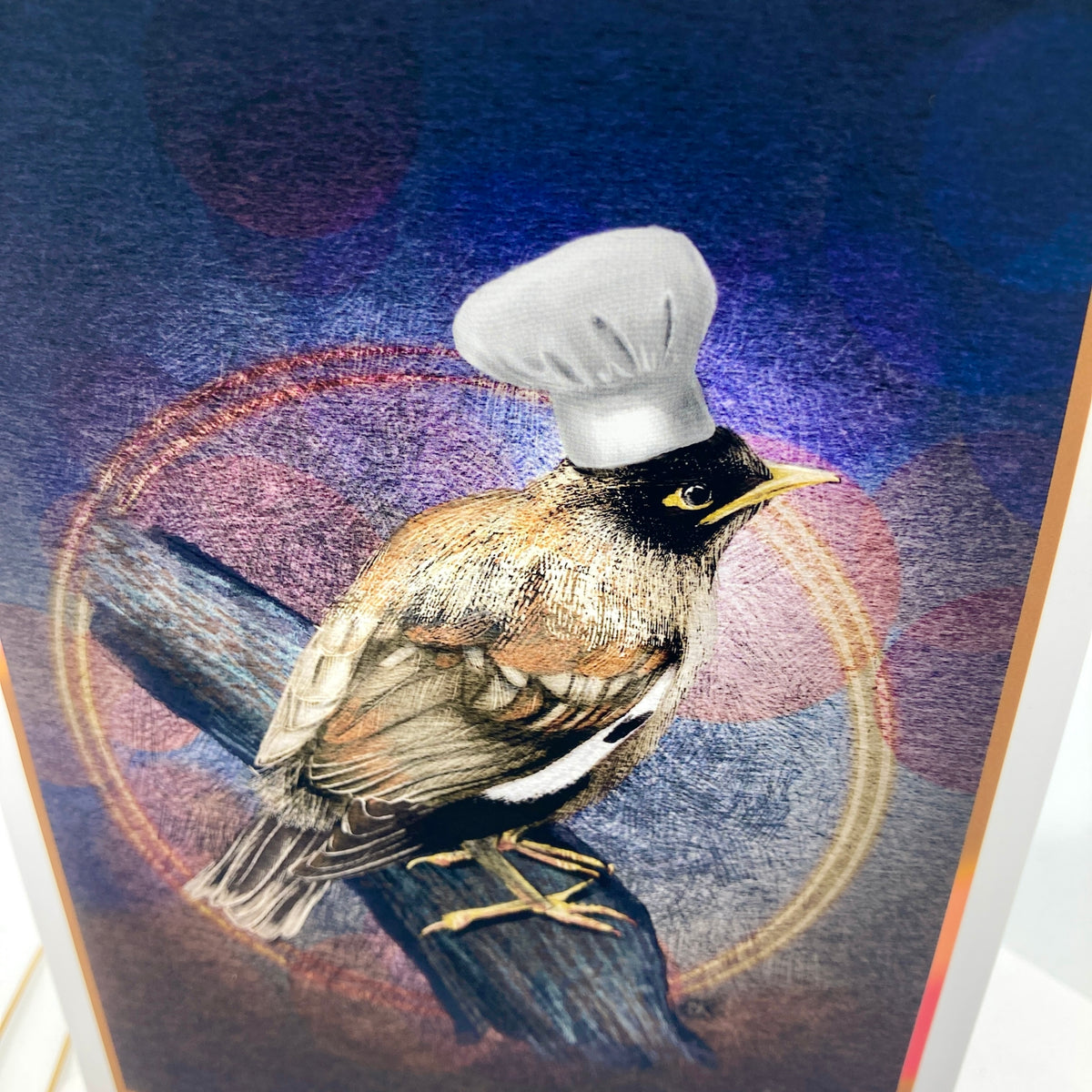 A close-up of a colorful art print or card featuring a bird perched on a branch, wearing a white chef's hat, against a blue and purple abstract background.
