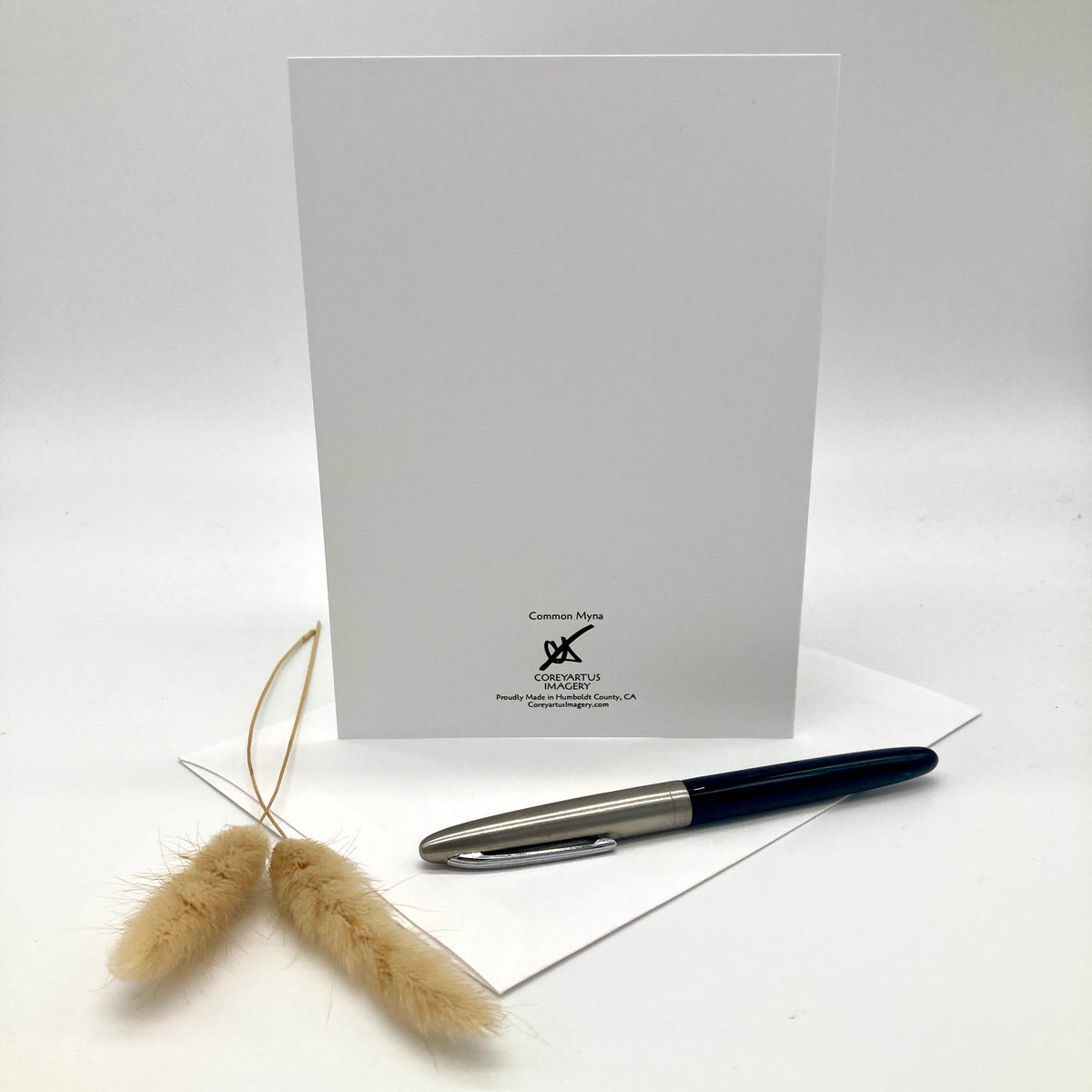 A white stationery card with black printed text and logo at the bottom, displayed upright on a white surface with a silver and black pen resting on top and dried beige grass stems in the foreground.