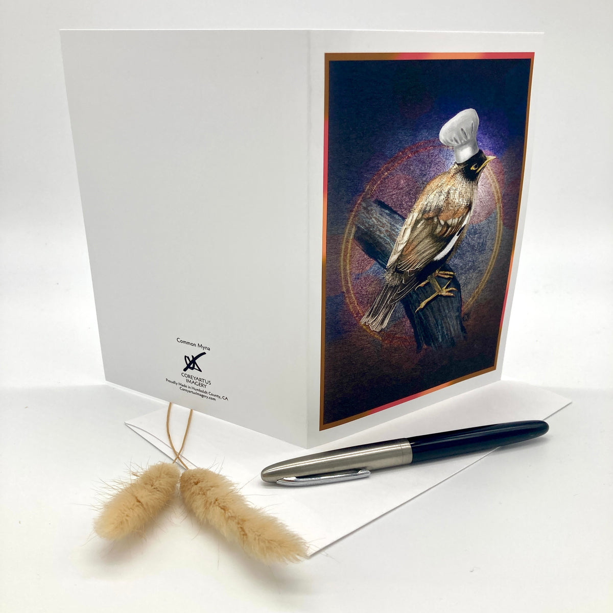 A white greeting card standing open on a white surface, featuring a printed illustration of a bird wearing a small white chef's hat on the front panel. A black and silver pen and a small beige decorative grass stem rest in front of the card.