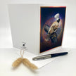 A white greeting card standing open on a white surface, featuring a printed illustration of a bird wearing a small white chef's hat on the front panel. A black and silver pen and a small beige decorative grass stem rest in front of the card.