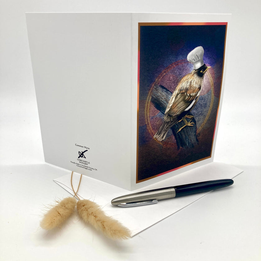 A white greeting card standing open on a white surface, featuring a printed illustration of a bird wearing a small white chef's hat on the front panel. A black and silver pen and a small beige decorative grass stem rest in front of the card.
