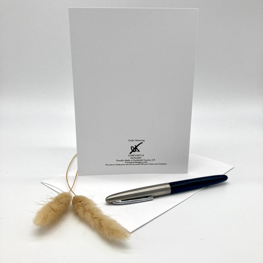 A white square card or envelope standing upright on a white surface, with a black and silver pen resting on a white sheet in front and two dried beige grass stems to the left. Small printed branding is visible near the bottom center of the white square piece.