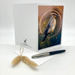 A white greeting card standing upright on a white surface, featuring an illustrated bird perched in front of a circular glowing motif. A matching white envelope lies in front, along with a black-and-silver pen and two small dried grass stems.