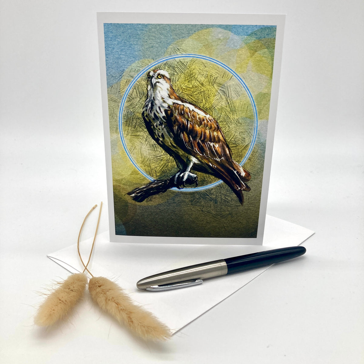 A vertical greeting card featuring a detailed illustration of an osprey perched against a textured blue and olive background, styled on a white surface with a pen, envelope, and dried bunny tail grass.