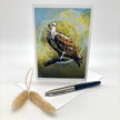 A vertical greeting card featuring a detailed illustration of an osprey perched against a textured blue and olive background, styled on a white surface with a pen, envelope, and dried bunny tail grass.