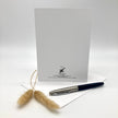A white stationery card with black printed text and logo stands upright on a white surface, with a silver and navy pen resting on a white envelope in front and dried beige grass stems to the left, against a clean white studio background.