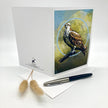 A white greeting card is shown open on a white surface, featuring an illustration of an osprey in a blue circular frame on the right panel. The left panel has small black printed text and logo near the bottom.