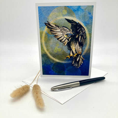 A vertical greeting card featuring a detailed illustration of a hawk with wings spread inside a circular frame, set against a blue and green textured background. The card is displayed standing upright on a white surface next to a black and silver pen, a white envelope, and two dried bunny tail grass stems.