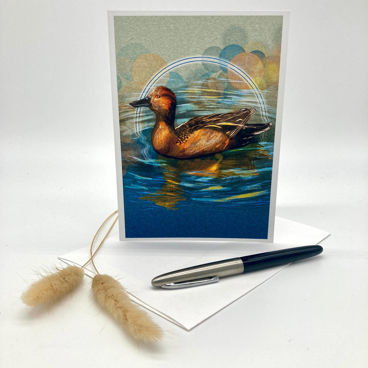 A vertical art print or greeting card featuring a detailed illustration of a brown duck swimming in blue water, styled on a white surface with a white envelope, a black and silver pen, and two dried beige grass stems.