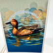 A vertical art print or card featuring a painted duck swimming in blue water, with abstract circular bokeh-like shapes in muted blue, yellow, and beige tones behind it. The print is photographed standing against a white background.