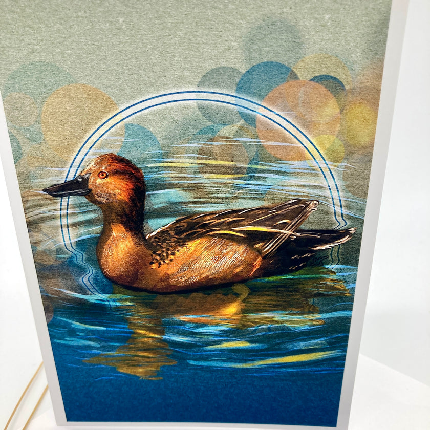 A vertical art print or card featuring a painted duck swimming in blue water, with abstract circular bokeh-like shapes in muted blue, yellow, and beige tones behind it. The print is photographed standing against a white background.