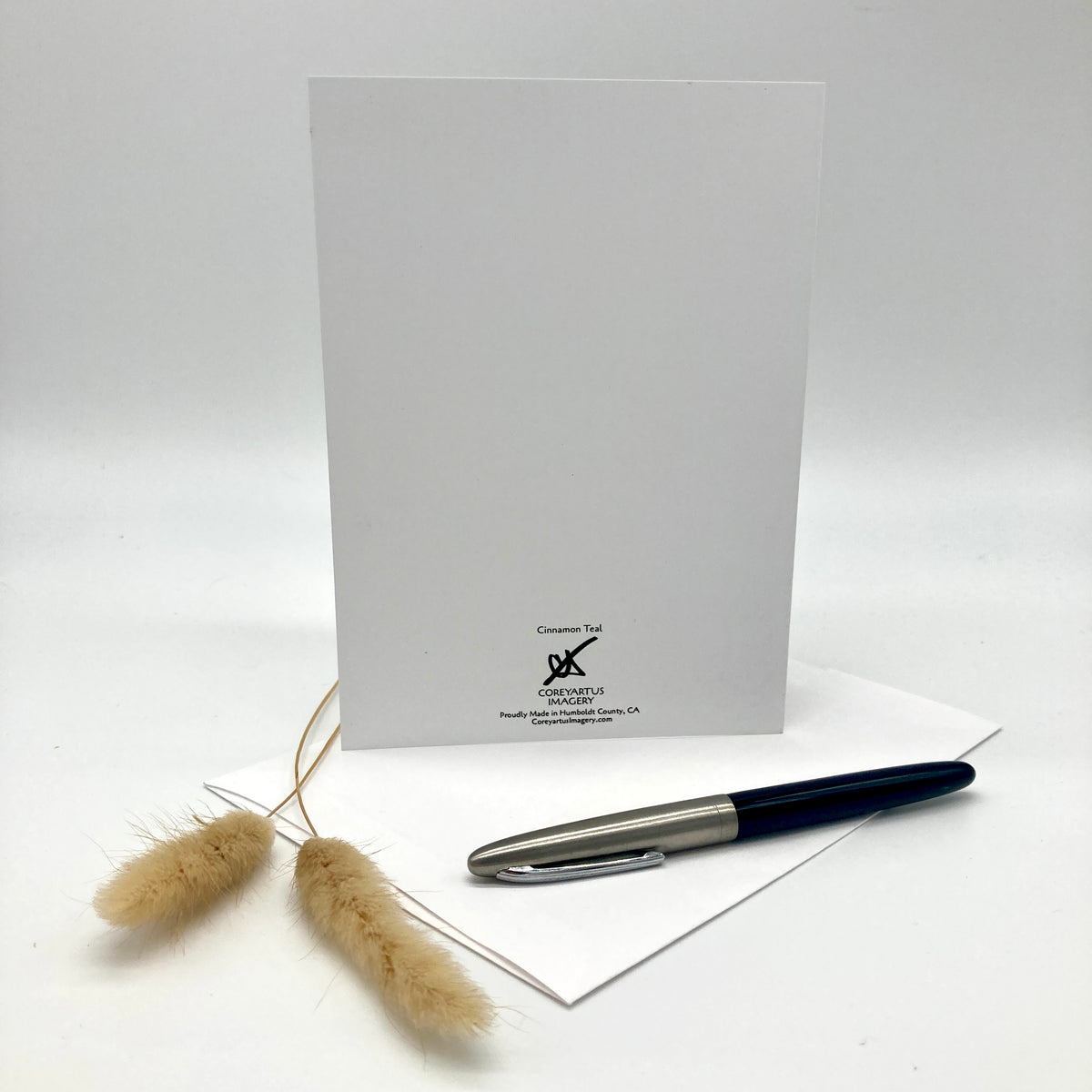 A white greeting card with a black printed logo at the bottom, displayed upright with a white envelope and a silver and black pen on a white surface. Two dried beige grass stems sit to the left of the card.