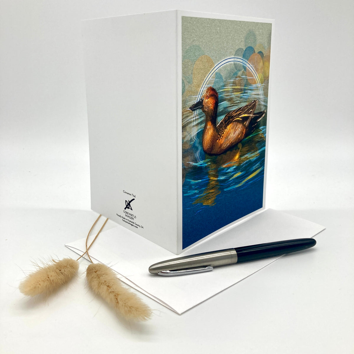 A white greeting card standing upright on a white surface, featuring a printed illustration of a brown duck in blue water on the front panel. A matching white envelope and a black-and-silver pen are placed in front, with two small dried grass stems to the left.
