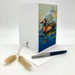 A white greeting card standing upright on a white surface, featuring a printed illustration of a brown duck in blue water on the front panel. A matching white envelope and a black-and-silver pen are placed in front, with two small dried grass stems to the left.