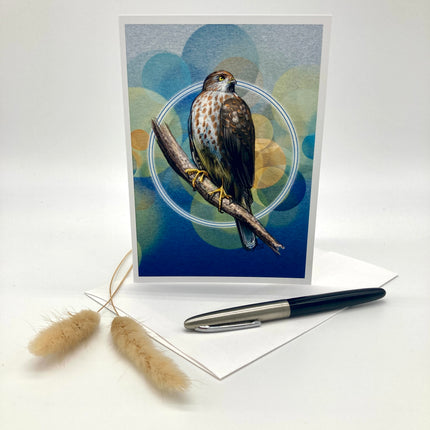 A greeting card featuring a detailed illustration of a bird of prey perched on a branch against a blue and teal abstract background, displayed upright on a white surface with an envelope, a black and silver pen, and two dried grass stems.