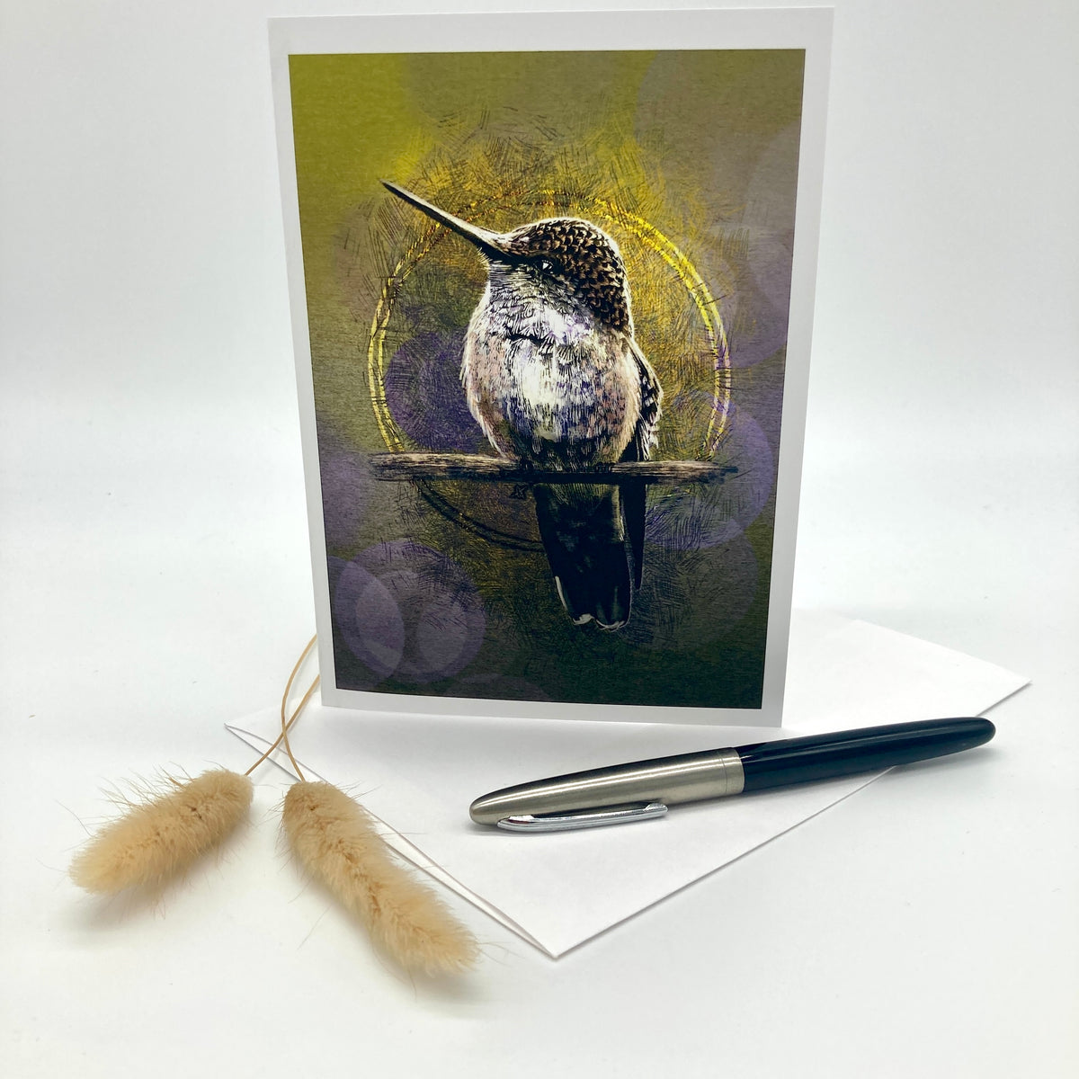 A vertical greeting card featuring a detailed illustration of a hummingbird perched on a branch, displayed on a white surface with a white envelope, a black and silver pen, and two dried bunny tail grasses.