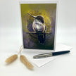 A vertical greeting card featuring a detailed illustration of a hummingbird perched on a branch, displayed on a white surface with a white envelope, a black and silver pen, and two dried bunny tail grasses.