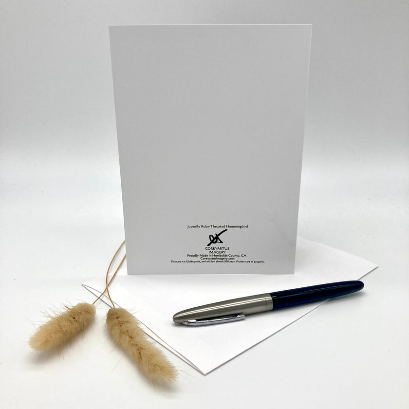 A white stationery card with black printed text and logo stands upright on a white surface, with a silver and navy pen resting on a white envelope in front and two dried bunny tail grass stems to the left. The card is branded 'Jovellita Kelly Florist Arrangement' and 'Pleasanton, CA'.