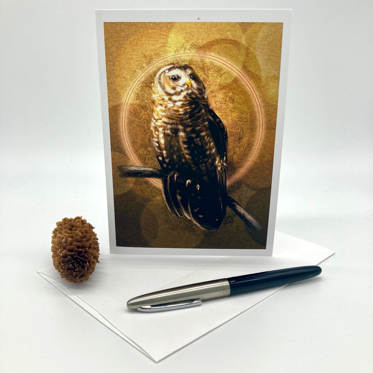 A vertical greeting card featuring a detailed illustration of an owl perched against a warm golden circular background, displayed on a white envelope with a black pen and a small pinecone prop on a white studio background.