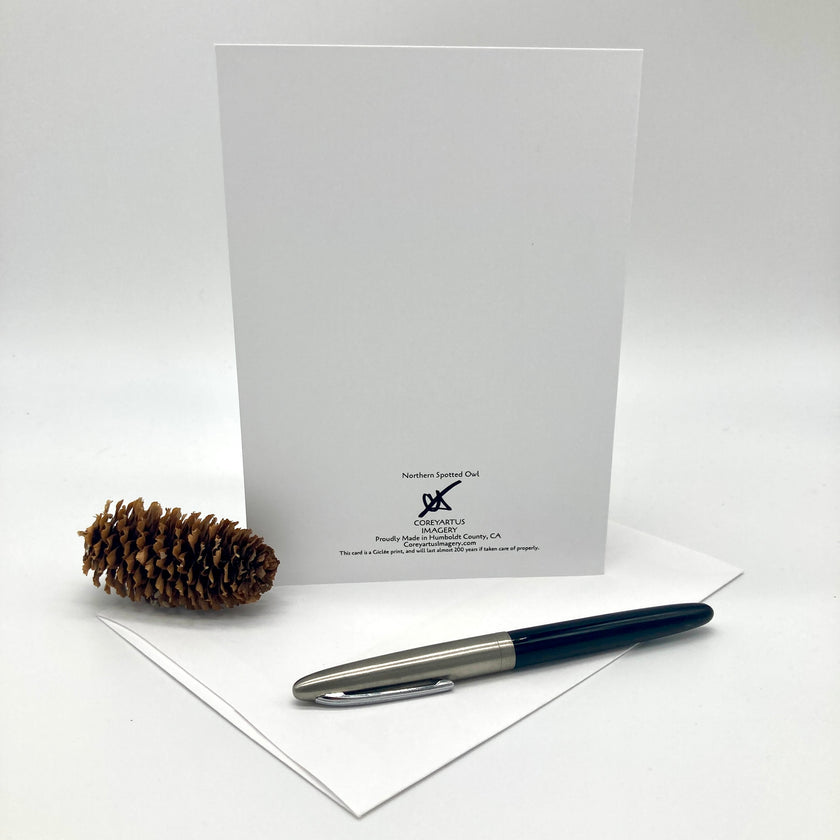 A white greeting card with a small black printed logo and text at the bottom, displayed upright on a white surface with an envelope, a black and silver pen, and a pinecone prop against a light gray studio background.