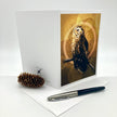 A white greeting card standing upright on a white surface, printed with a detailed owl perched against a warm gold-toned circular background. A pinecone and a black-and-silver pen are placed in front of the card.