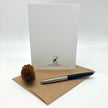 A white card with the 'Proud Father' logo and address printed at the bottom, displayed upright on a brown kraft envelope with a silver and navy blue pen and a small pinecone on a white background.